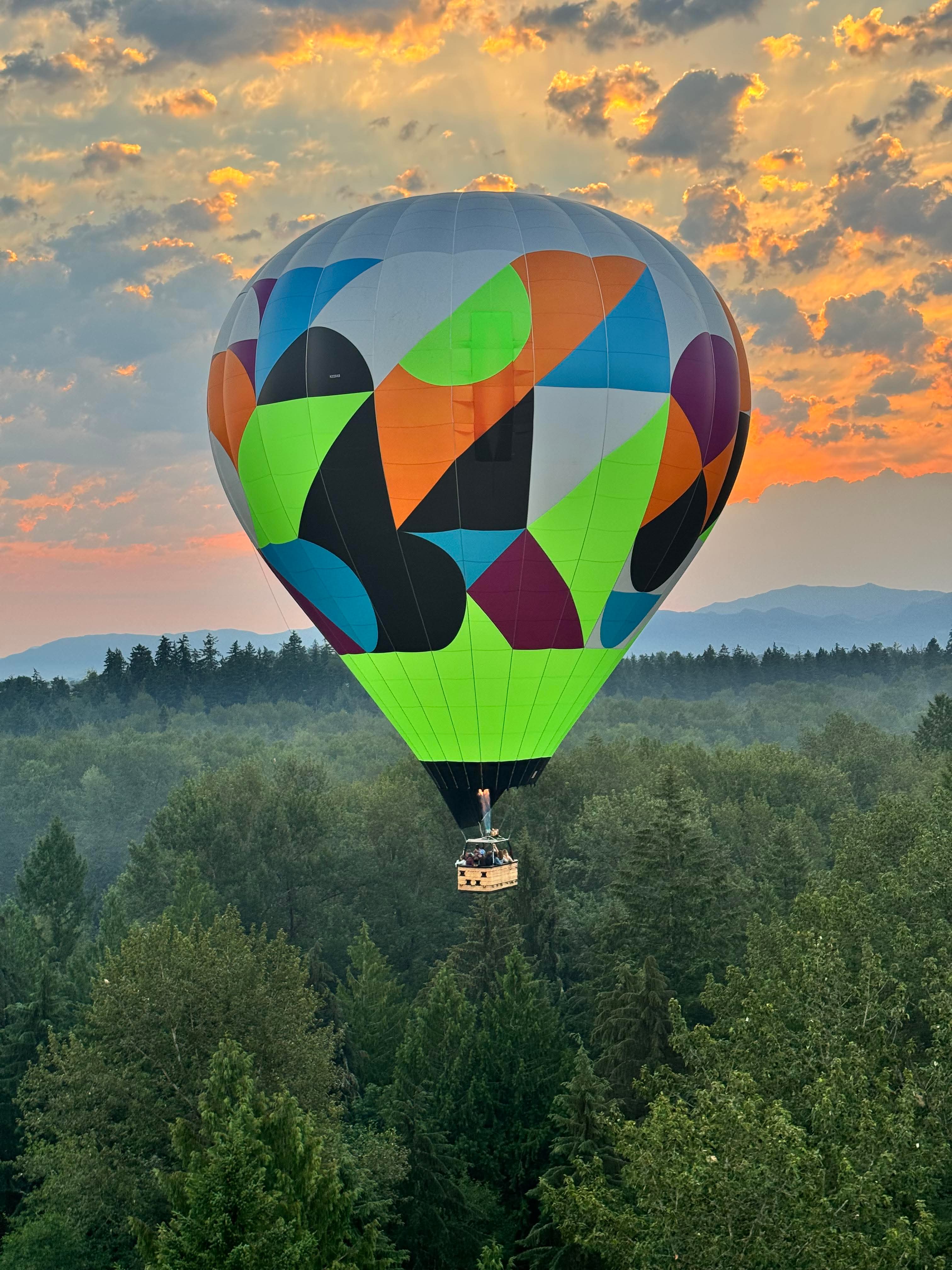 Colorful hot air balloon flying over lush green forest with mountains and golden sunset sky